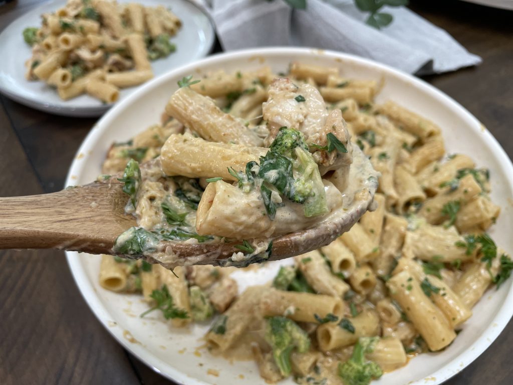 A bowl of pasta, chicken, broccoli and spinach, scooped with a wooden spoon.