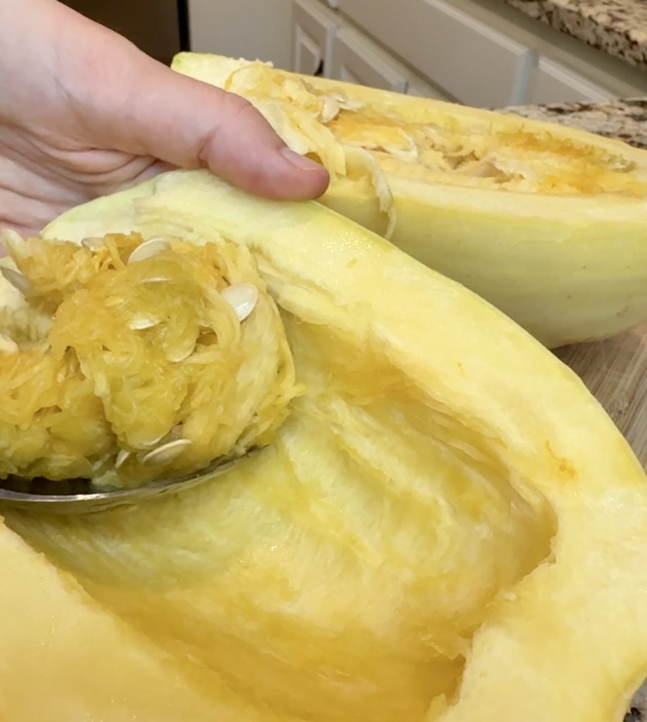 A hand uses a spoon to scoop out seeds and pulp from a halved raw spaghetti squash on a kitchen counter, prepping it for a delicious low-carb dinner or baked feta recipe.