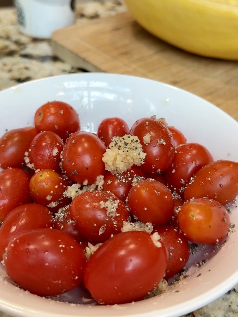 A white bowl filled with grape tomatoes, topped with minced garlic and herbs, sits on a kitchen counter—perfect as a topping for spaghetti squash in a delicious low-carb dinner. A cutting board and yellow squash are visible in the background.