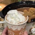 Hot cocoa in a paper mug, topped with a cookie and a slow cooker in the back