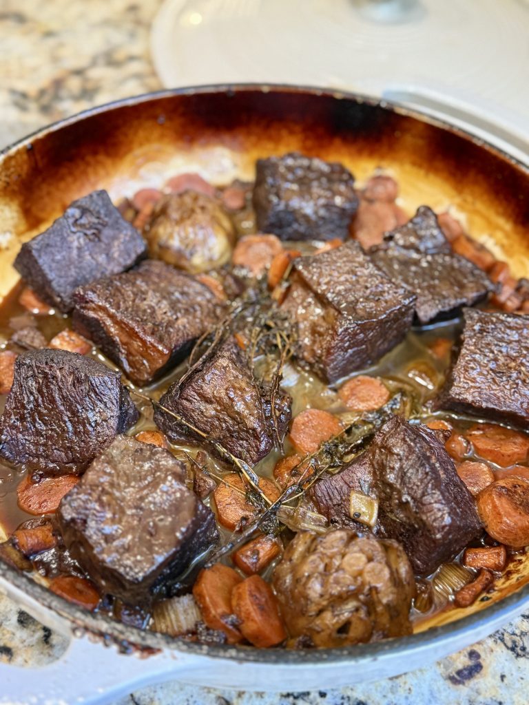 A pot of slow-cooked beef with large chunks of meat, sliced carrots, whole garlic, fresh herbs, and broth—all simmered together in a round, enameled Dutch oven on a granite countertop.