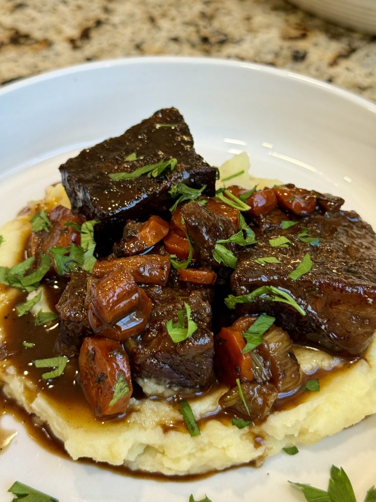 Slow-cooked beef short ribs with carrots in a rich brown sauce, served over creamy mashed potatoes and garnished with fresh parsley—a comforting crockpot dinner presented on a white plate.