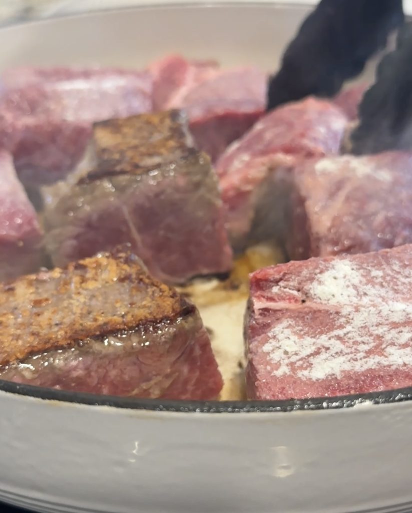 Large chunks of chuck roast are being seared in a white pan, with some pieces browned and others still raw. Black tongs grip one piece as steam rises, starting the first step in making tender red wine braised beef.