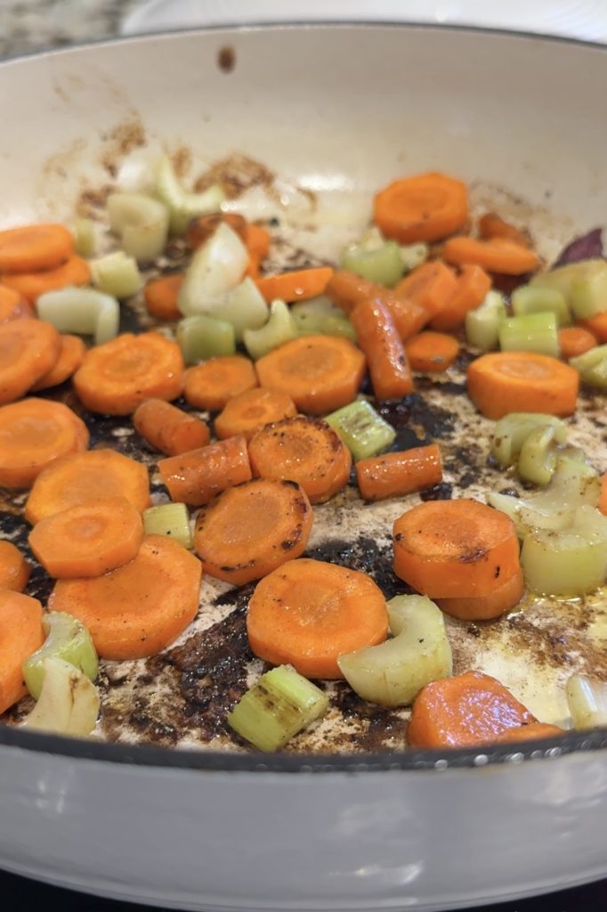 Sliced carrots and celery pieces sautéing in a white skillet, with browned bits and seasoning from chuck roast on the pan’s surface.