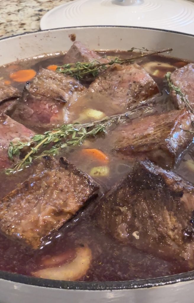Chunks of red wine braised beef, carrots, and garlic simmer in a rich brown broth, garnished with sprigs of fresh thyme in a large white pot.