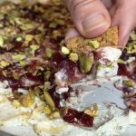A plate with Boursin cheese spread on it, topped with cranberry sauce and sprinkled with pistachios and a hand scooping the dip with a cracker