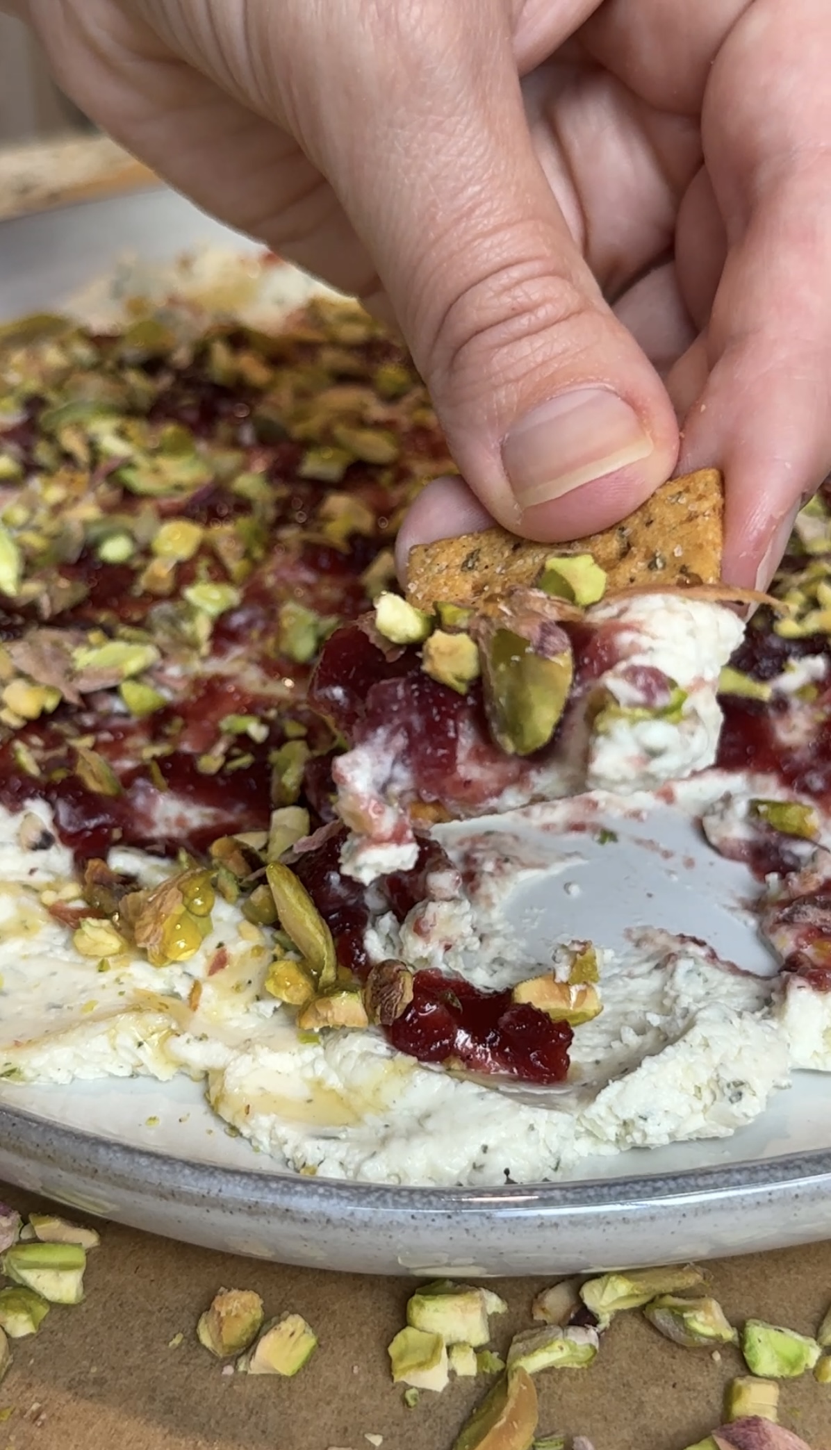 A plate with Boursin cheese spread on it, topped with cranberry sauce and sprinkled with pistachios and a hand scooping the dip with a cracker