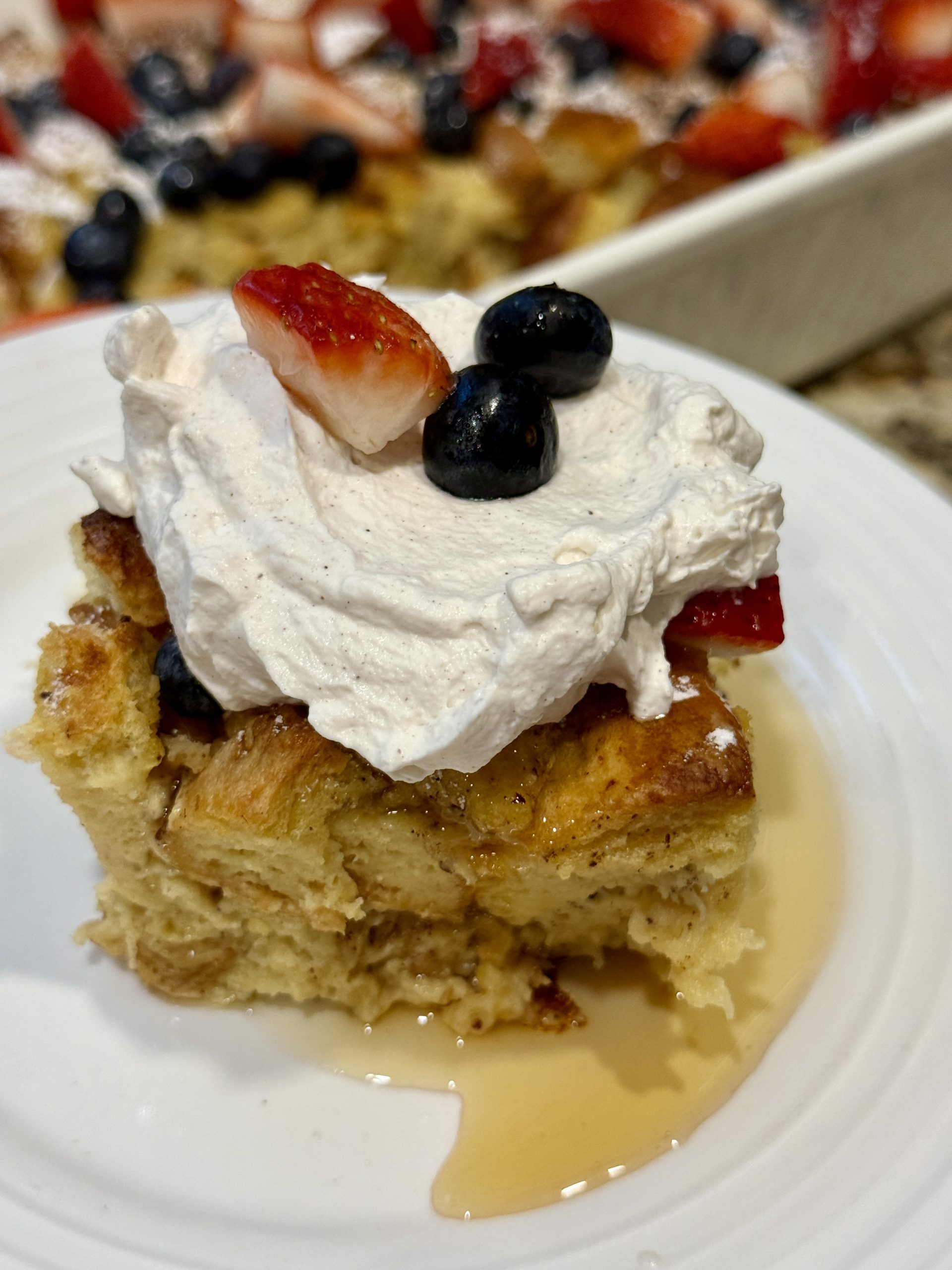A slice of French toast casserole topped with whipped cream, strawberries, and blueberries on a white plate. Syrup drizzles around the slice.