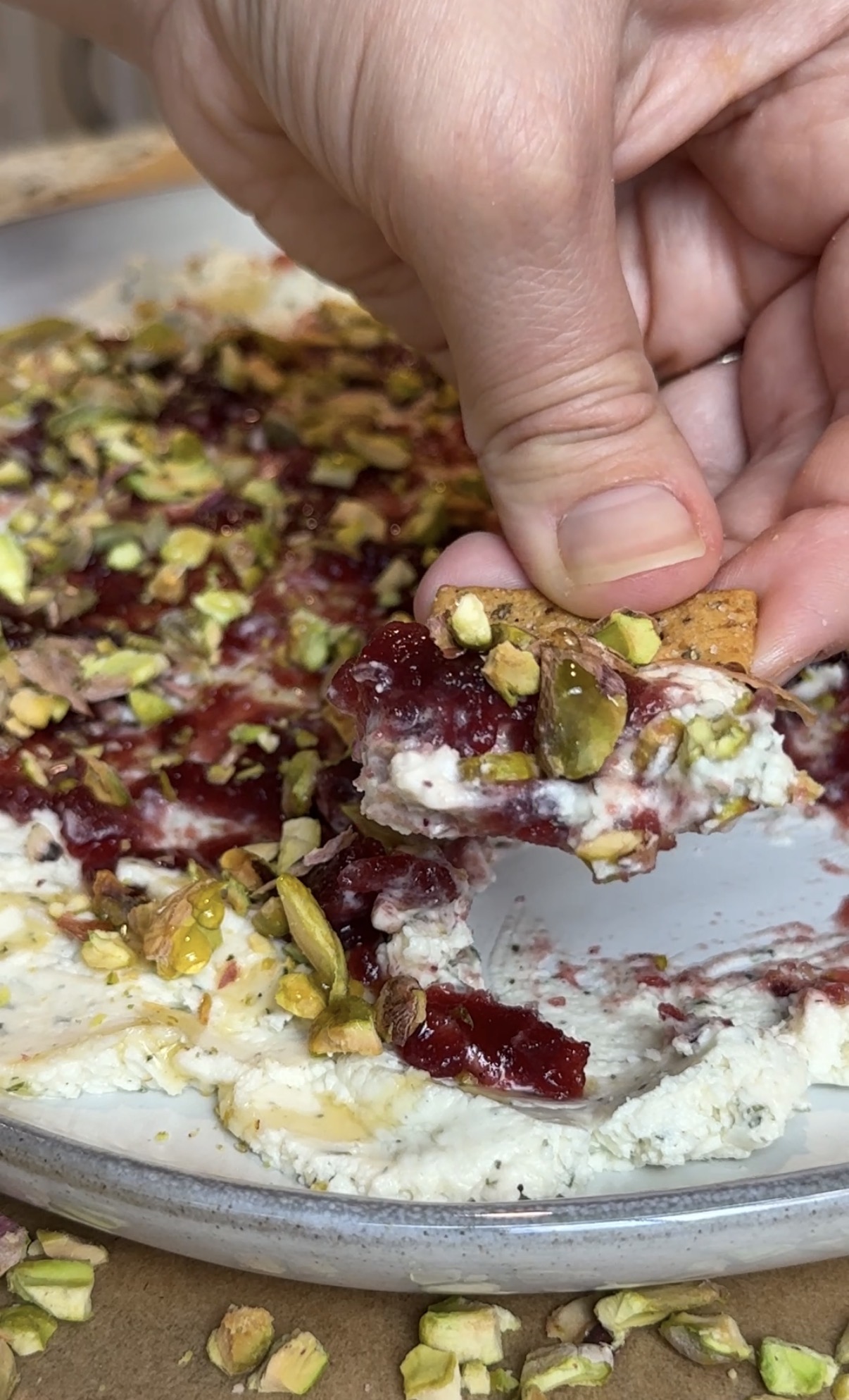 A serving platter with Boursin cheese, jam and pistachios and a person scooping the dip with a cracker.