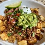 A bowl with crispy air fried potatoes, topped with shredded cheese, taco ground beef, guacamole, pico de gallo, sour cream, and a wedge of lime.