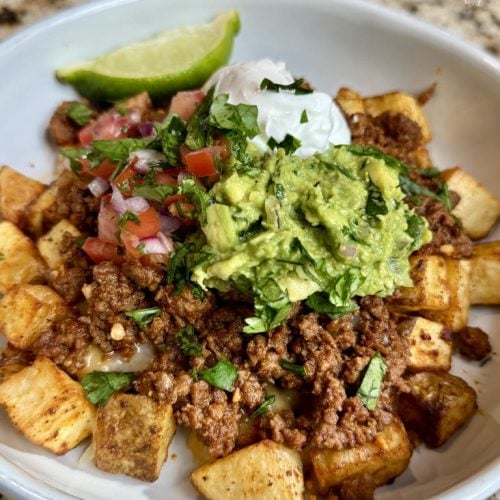 A bowl with crispy air fried potatoes, topped with shredded cheese, taco ground beef, guacamole, pico de gallo, sour cream, and a wedge of lime.