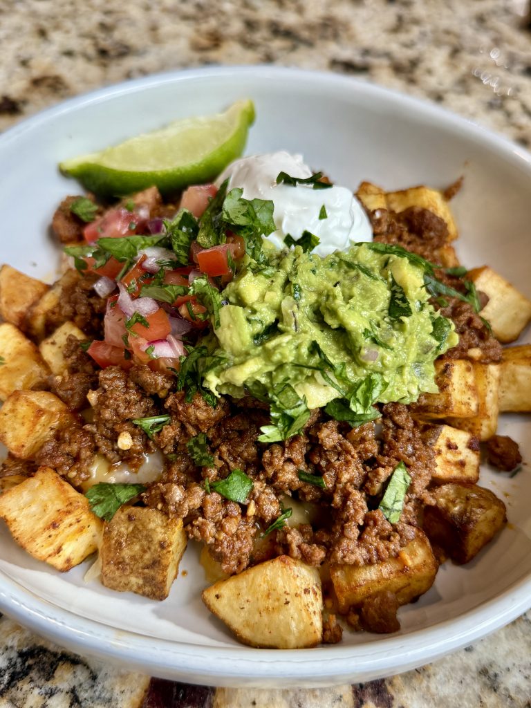 A bowl with crispy air fried potatoes, topped with shredded cheese, taco ground beef, guacamole, pico de gallo, sour cream, and a wedge of lime.
