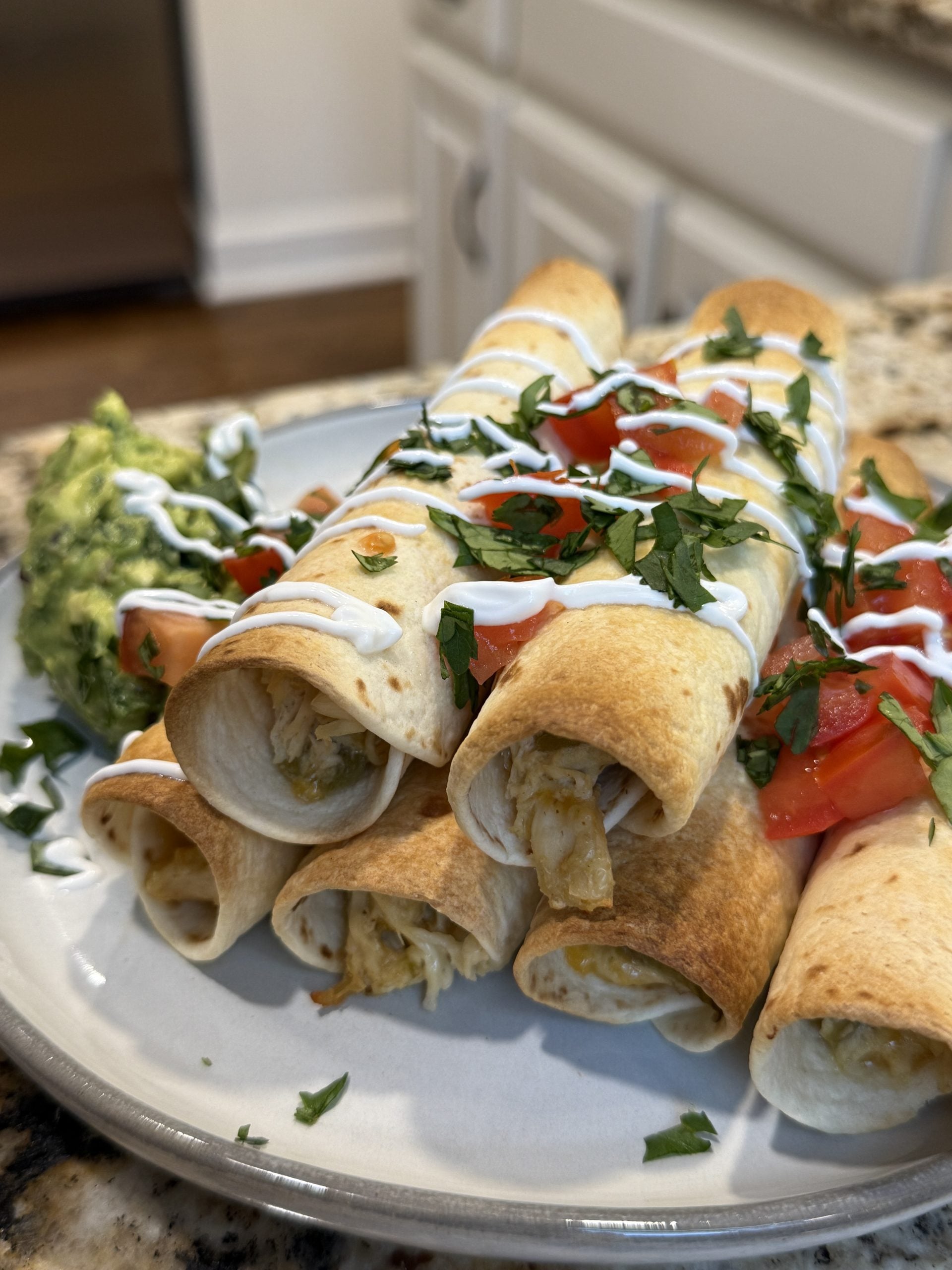 Crispy baked chicken taquitos piled on a white plate, topped with sour cream drizzle, diced tomatoes, and fresh cilantro, served alongside a scoop of guacamole
