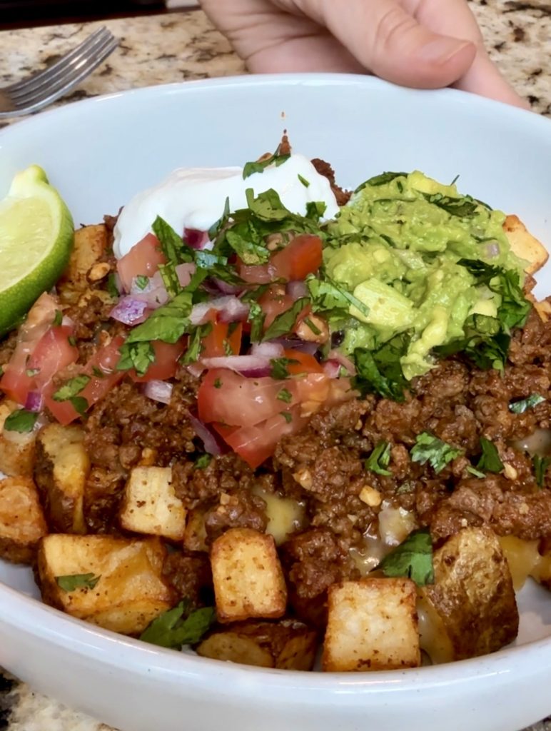 A bowl with crispy air fried potatoes, topped with shredded cheese, taco ground beef, guacamole, pico de gallo, sour cream, and a wedge of lime.