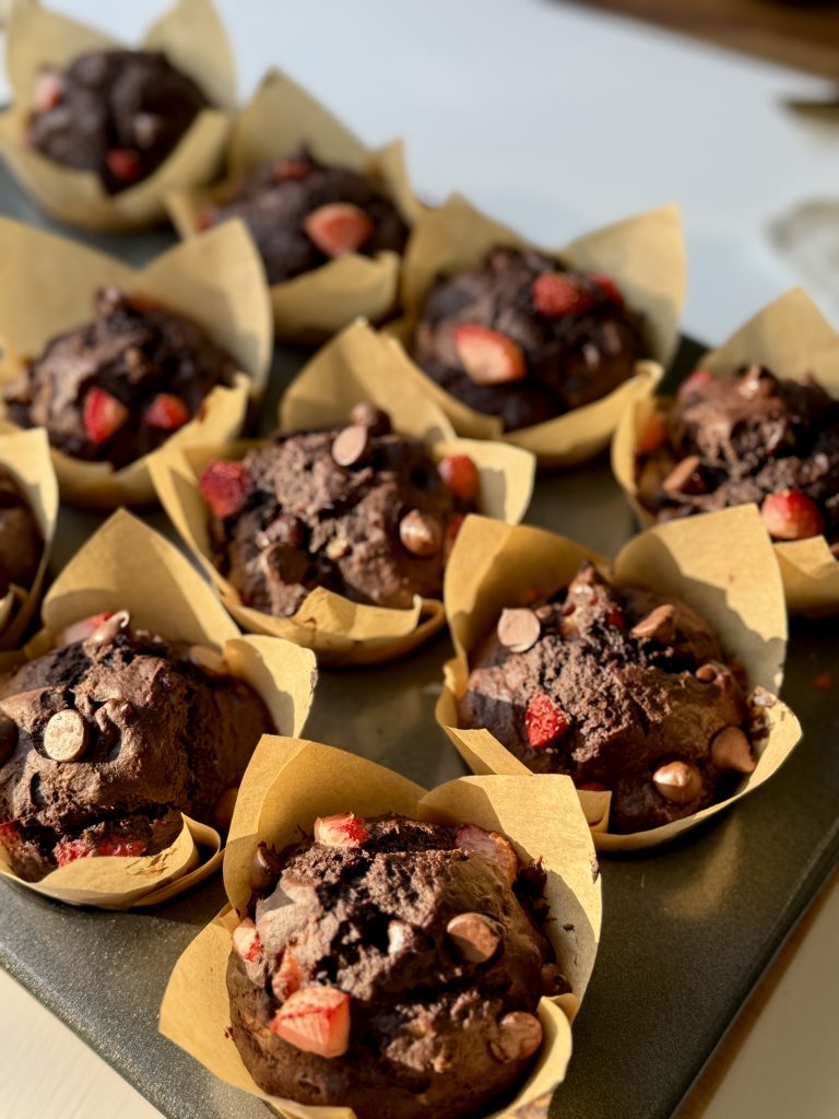 Twelve chocolate muffins in brown paper liners are topped with chocolate chips and strawberry pieces, placed in a muffin tray and lit by natural light.
