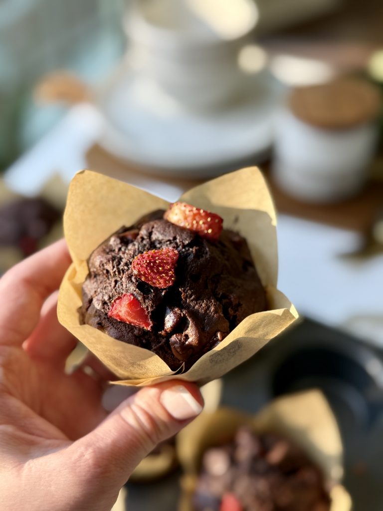 Hand holding a chocolate muffin in brown paper, topped with fresh strawberries. Soft natural light creates a warm and inviting atmosphere.