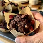 A picture of a hand holding a chocolate muffin with strawberries and chocolate chips. It's