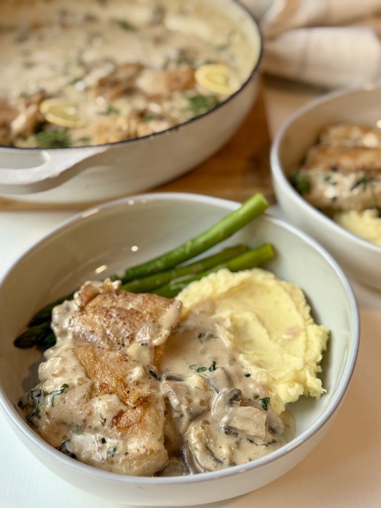 A bowl with creamy chicken, mushrooms, mashed potatoes, and green beans. In the background, a larger dish holds more of the same meal.