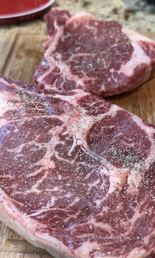 Two marbled raw steaks seasoned with black pepper sit on a wooden cutting board. The texture and rich marbling suggest a juicy, flavorful cut.