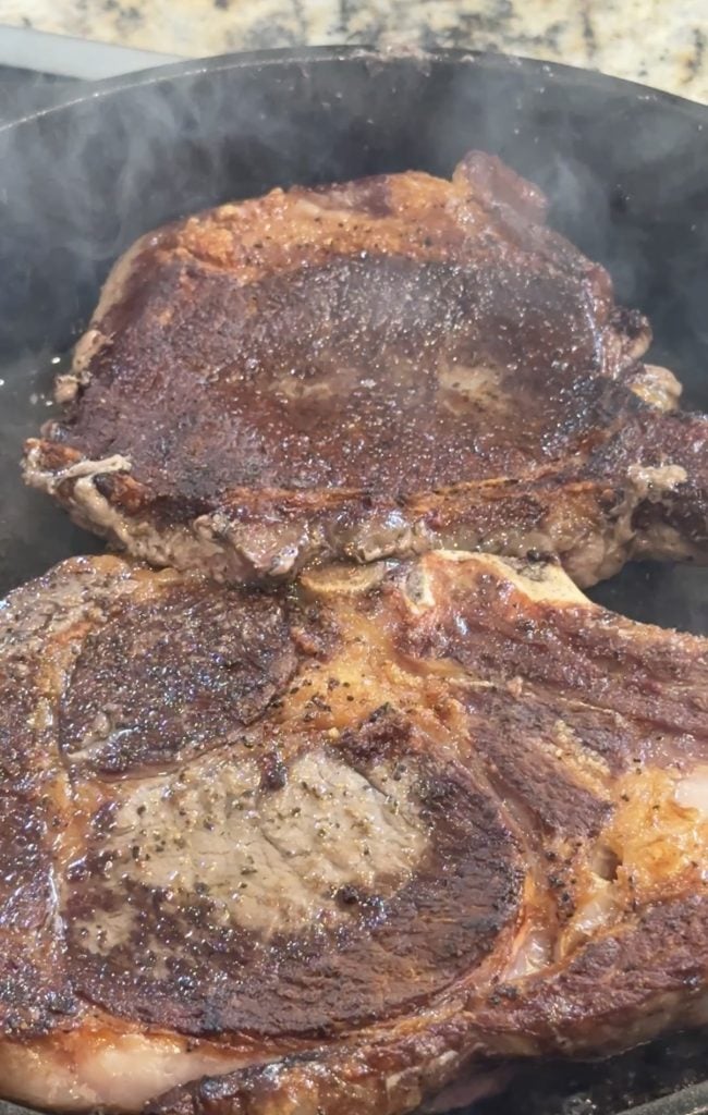 Two thick, juicy steaks sizzle in a pan, demonstrating a rich, browned crust. Steam rises, evoking a sense of warmth and savory aroma.