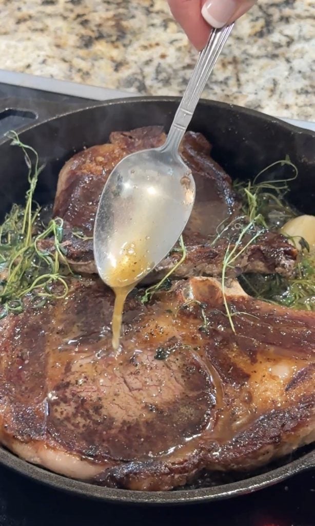 A juicy steak in a cast iron skillet is being basted with herb-infused butter using a spoon. Fresh thyme and garlic add aromatic flair.