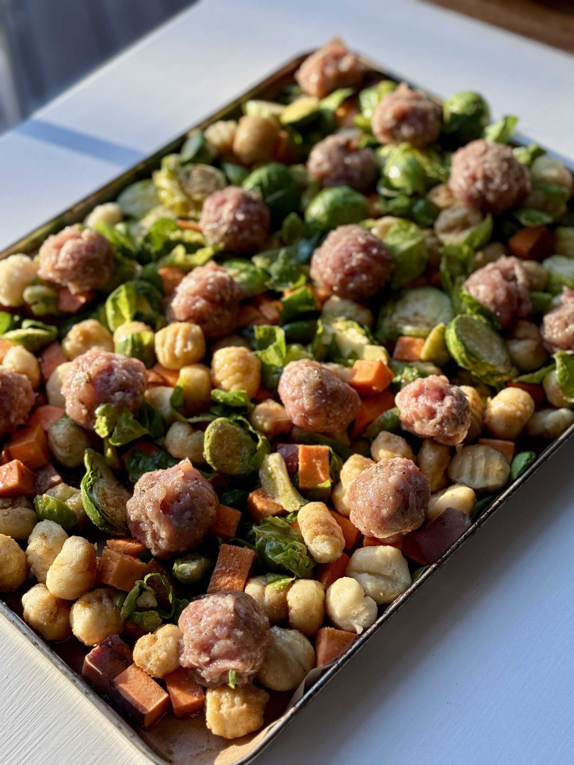 A baking sheet filled with raw meatballs, gnocchi, Brussels sprouts, and sweet potato cubes, bathed in warm sunlight, evoking a cozy, homey feel.