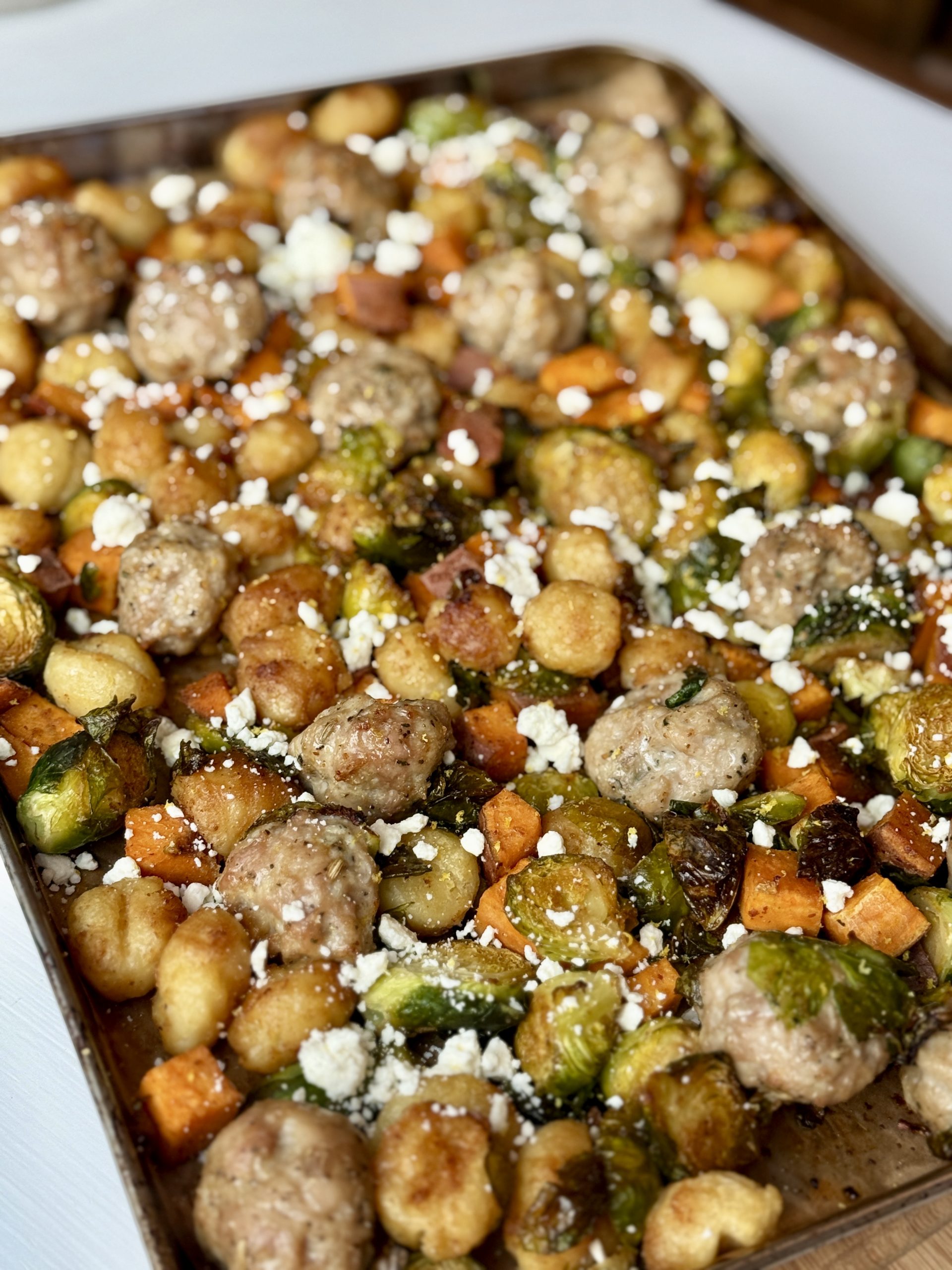 A sheet pan filled with roasted meatballs, Brussels sprouts, gnocchi, and diced sweet potatoes, topped with crumbled feta cheese, creating a hearty meal.