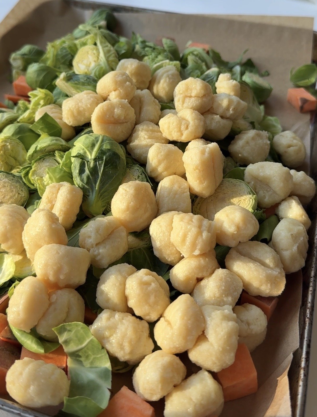 Close-up of a baking tray topped with uncooked gnocchi, halved Brussels sprouts, and diced sweet potatoes, ready for roasting. Natural lighting highlights textures.