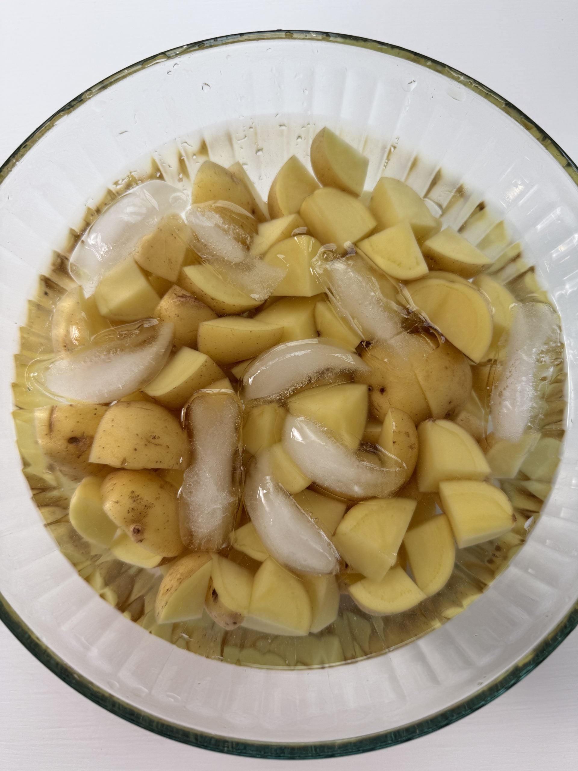 A glass bowl filled with quartered potatoes soaking in water with several ice cubes on top.