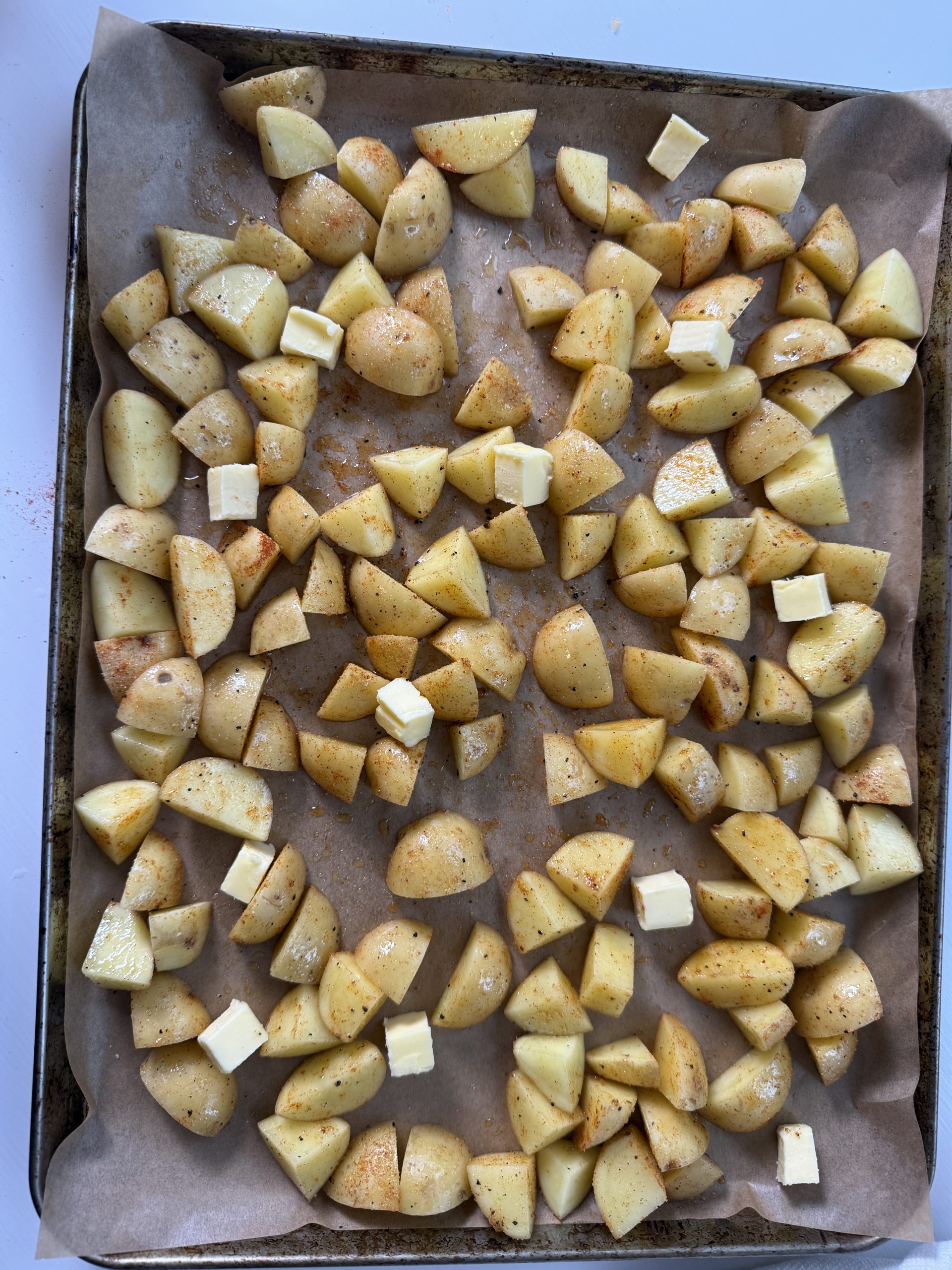 Overhead view of chopped yellow potatoes spread on a parchment-lined sheet pan, lightly seasoned and topped with small cubes of butter before roasting.