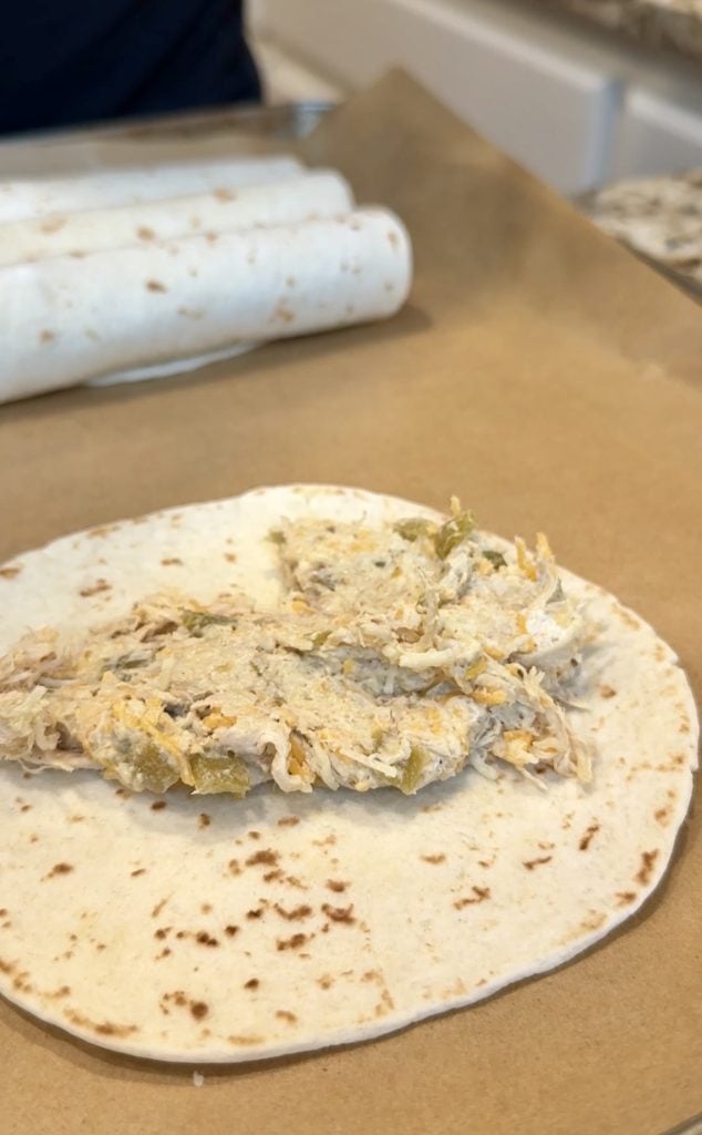 A flour tortilla topped with shredded chicken, cheese, and green chiles on parchment paper, ready to be rolled into taquitos, with already-rolled taquitos visible in the background