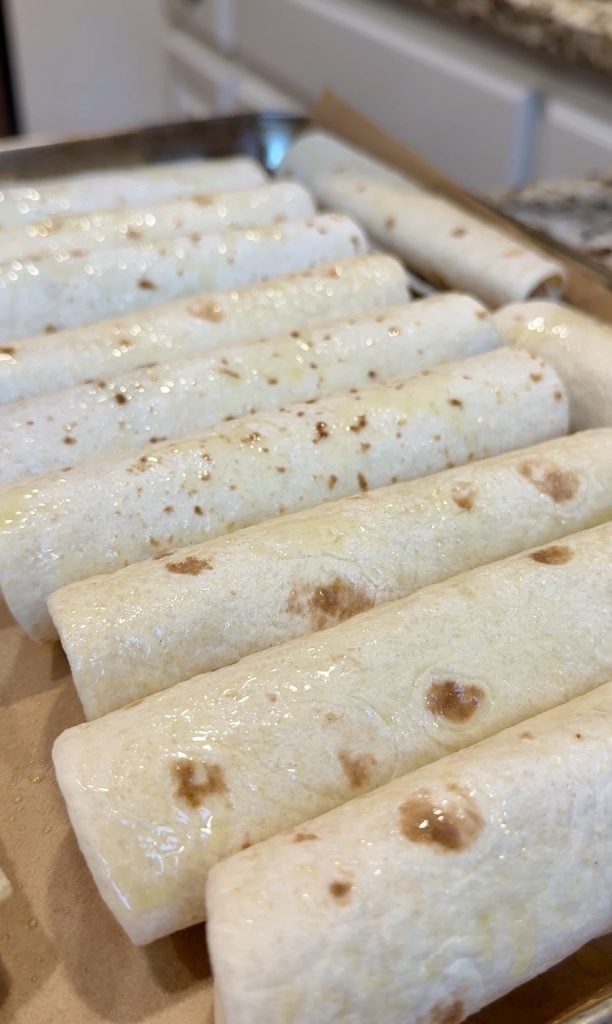 Uncooked flour tortilla taquitos brushed with oil and lined up on a parchment-lined baking sheet, ready to go into the oven