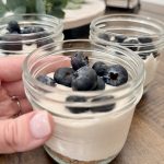 A hand holding a small glass jar filled with a layered dessert, topped with fresh blueberries. More jars with similar desserts are visible on a wooden table in the background.