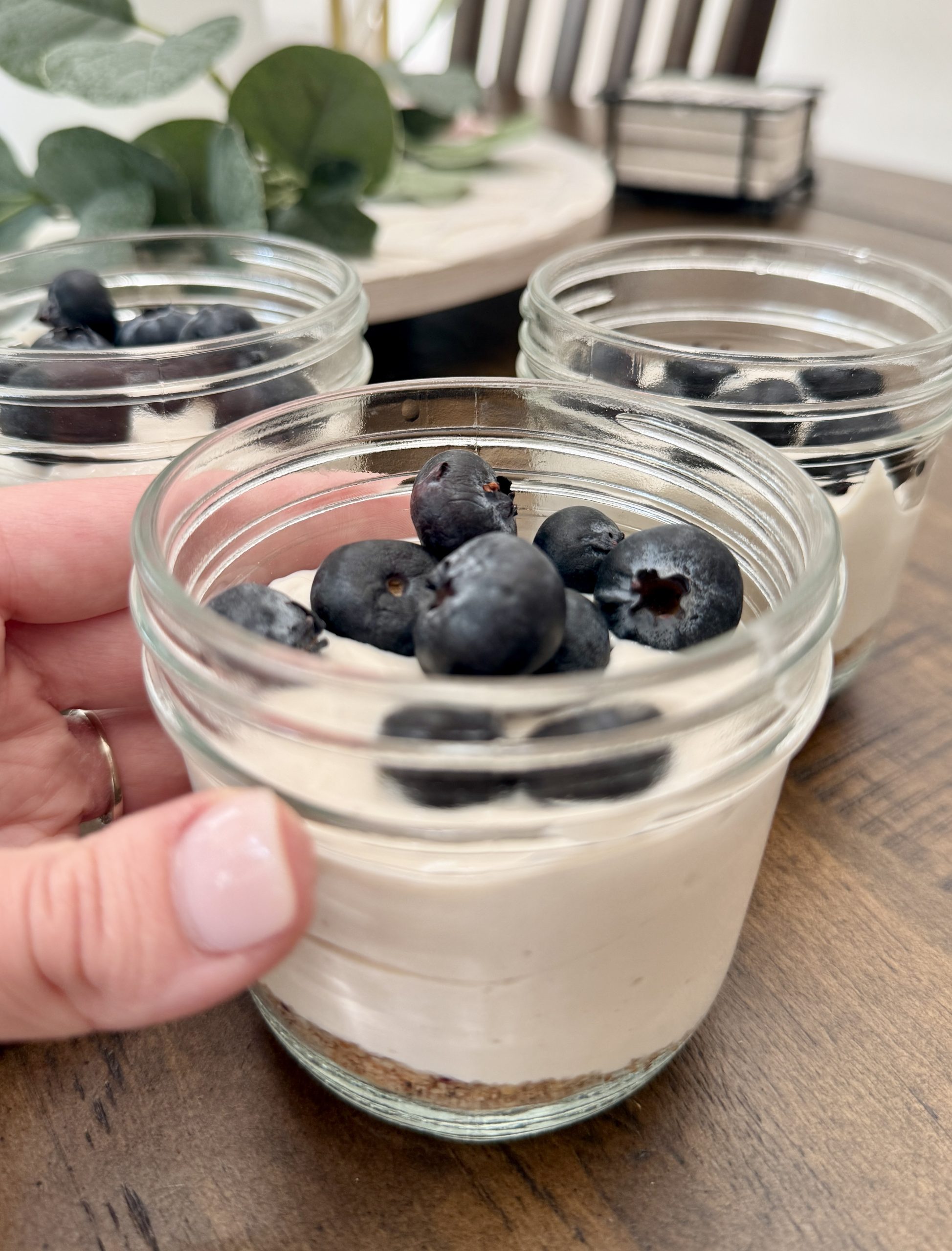 A hand holding a small glass jar filled with a layered dessert, topped with fresh blueberries. More jars with similar desserts are visible on a wooden table in the background.