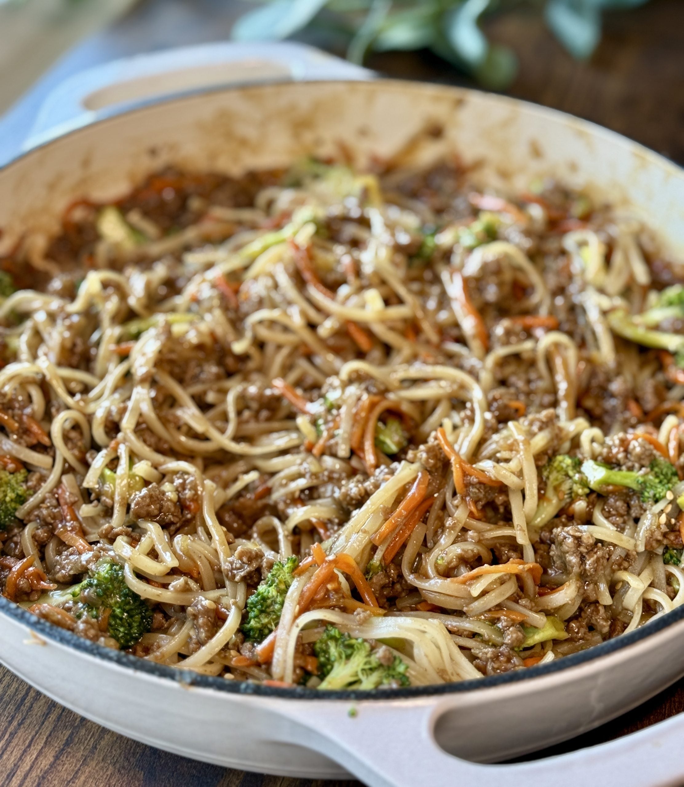 A skillet filled with stir-fried noodles, ground meat, broccoli, and shredded carrots in a savory sauce, sitting on a wooden table.