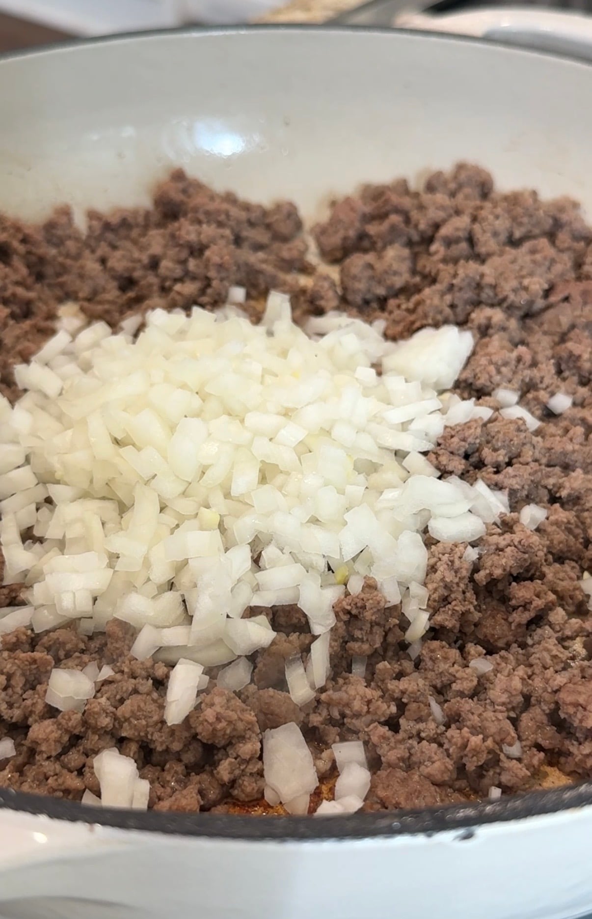 Chopped onions sit on top of browned ground beef in a white skillet, ready to be mixed and cooked together.