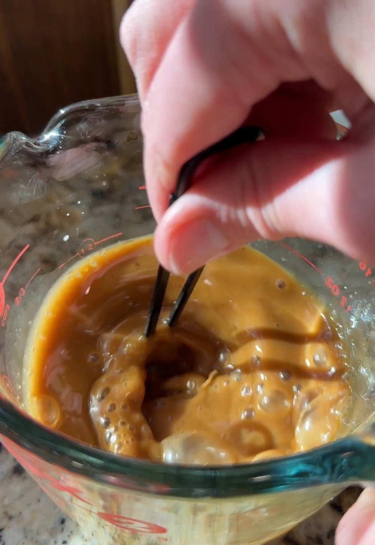 A hand uses a whisk to stir creamy brown liquid with bubbles in a glass measuring cup on a granite countertop.