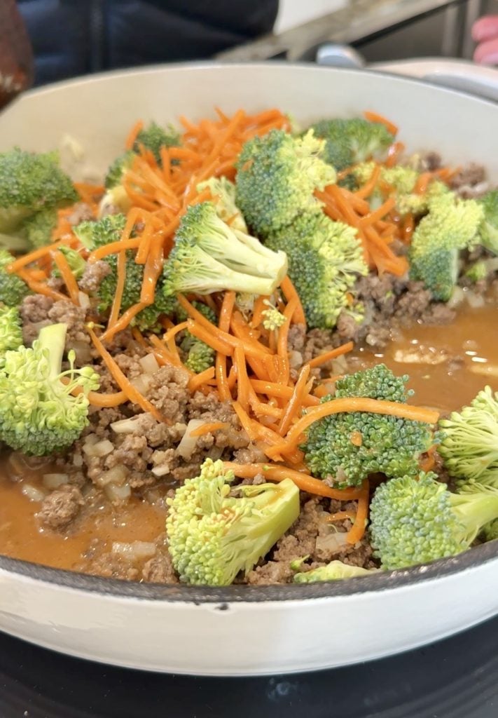 A close-up of a skillet filled with cooked ground beef, chopped onions, broccoli florets, and shredded carrots in a savory sauce.
