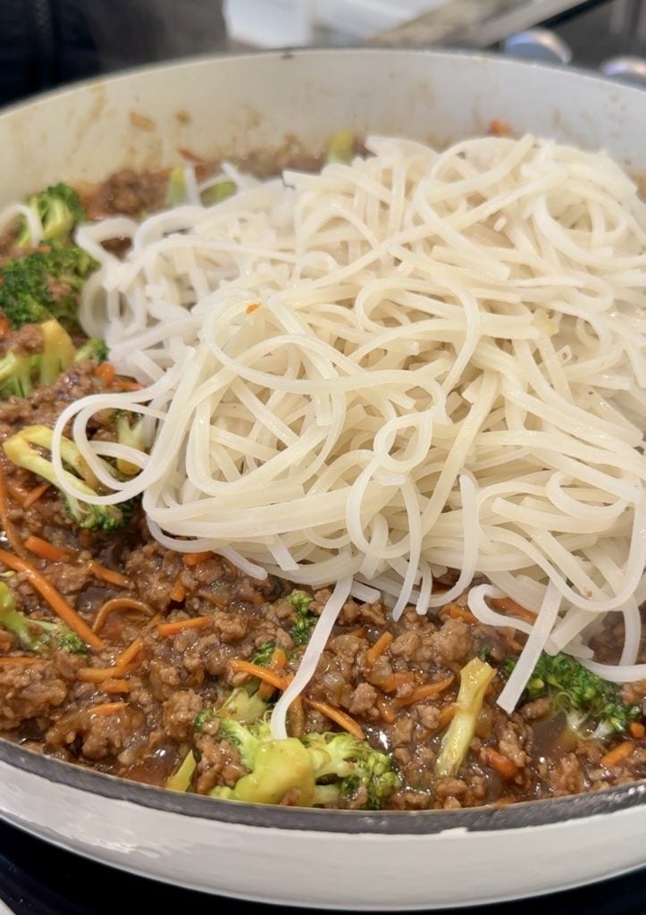 A close-up of a pan filled with cooked ground beef, broccoli, carrots, and a portion of cooked rice noodles on top, ready to be mixed together.