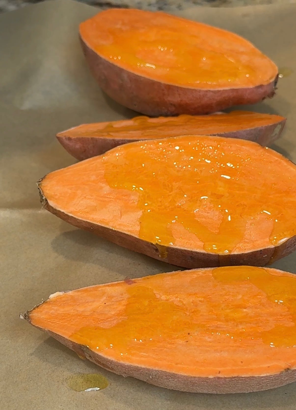 Four halved sweet potatoes with bright orange flesh sit on parchment paper, drizzled with honey or oil, ready for roasting.