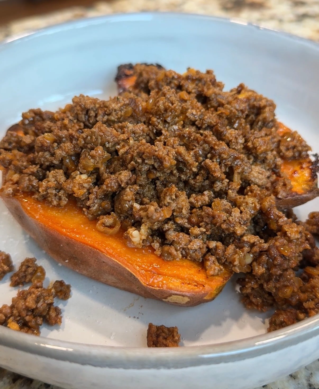 A baked sweet potato half topped with a generous portion of seasoned ground beef sits on a white plate. Some of the beef mixture has spilled onto the plate.