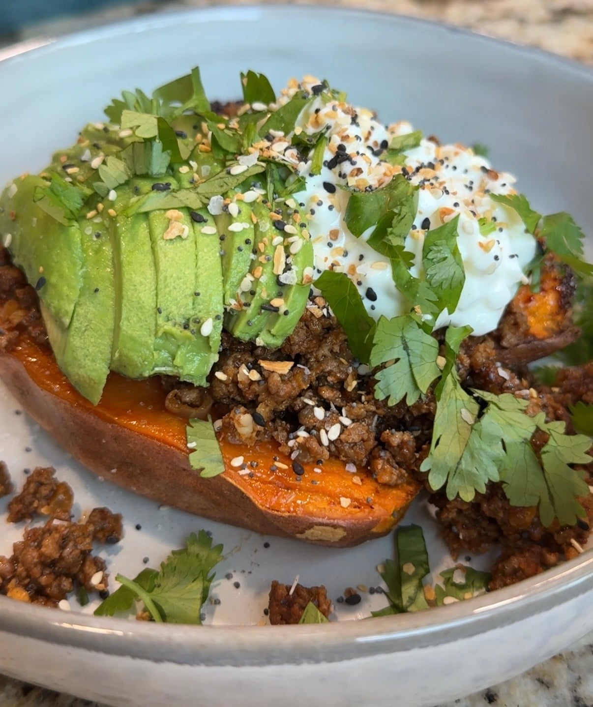 A baked sweet potato half topped with ground beef, sliced avocado, cottage cheese, chopped cilantro, and seasoning sits in a white bowl on a speckled countertop.