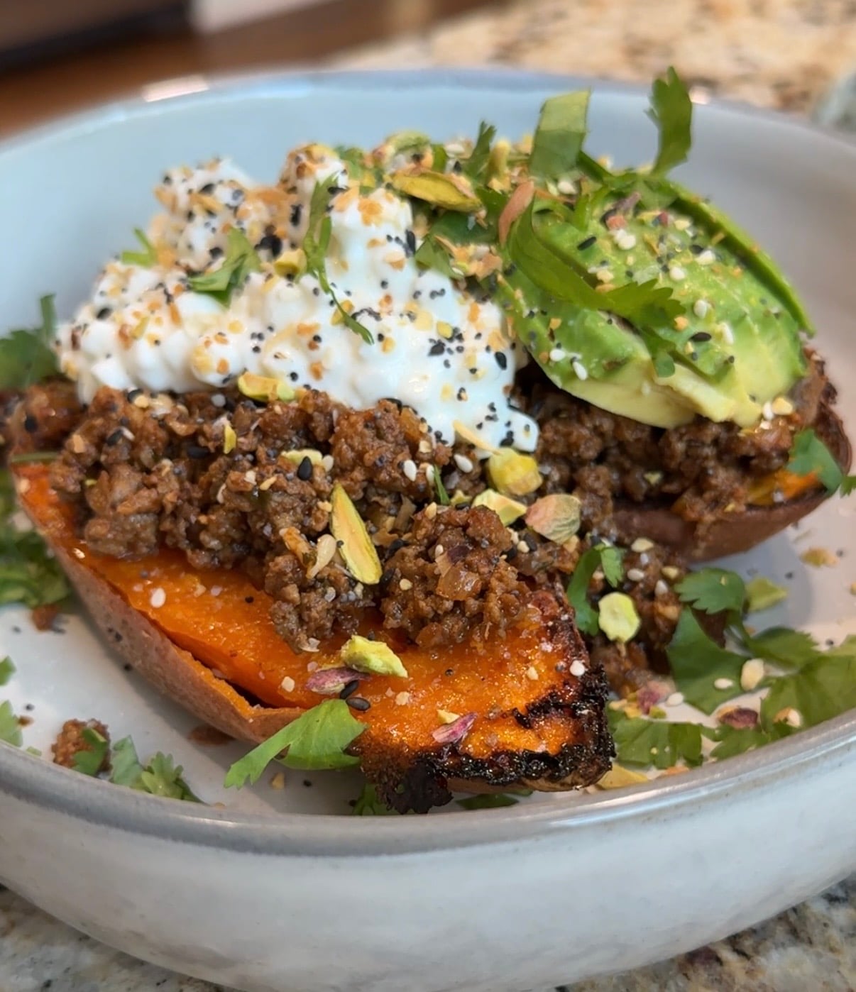 A halved roasted sweet potato filled with seasoned ground meat, topped with sliced avocado, cottage cheese, chopped pistachios, herbs, and seasoning, served in a shallow bowl on a granite countertop.