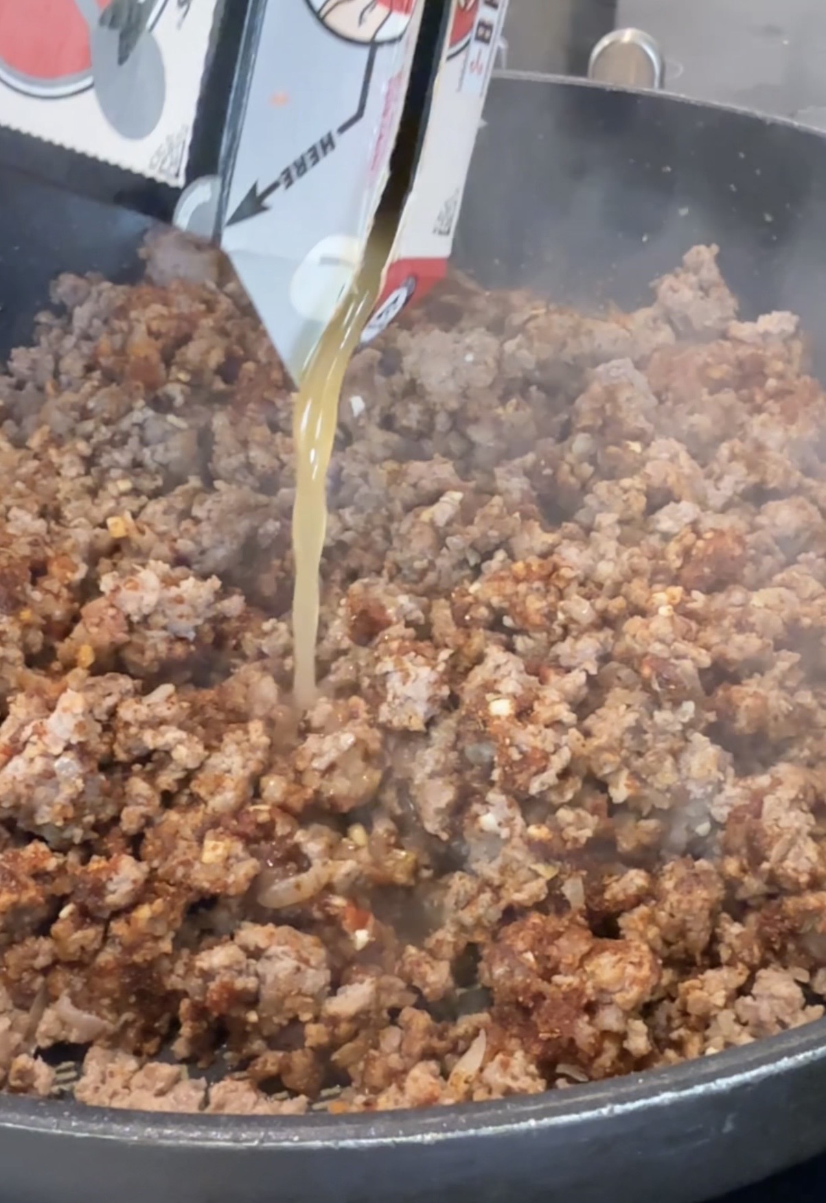 A close-up of cooked ground beef in a skillet as beef broth is being poured in from a carton, with steam rising from the pan.