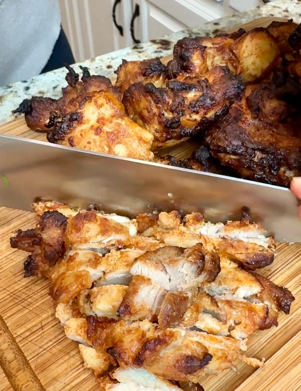 A wooden cutting board with cooked chicken thighs on it and a knife chopping the chicken