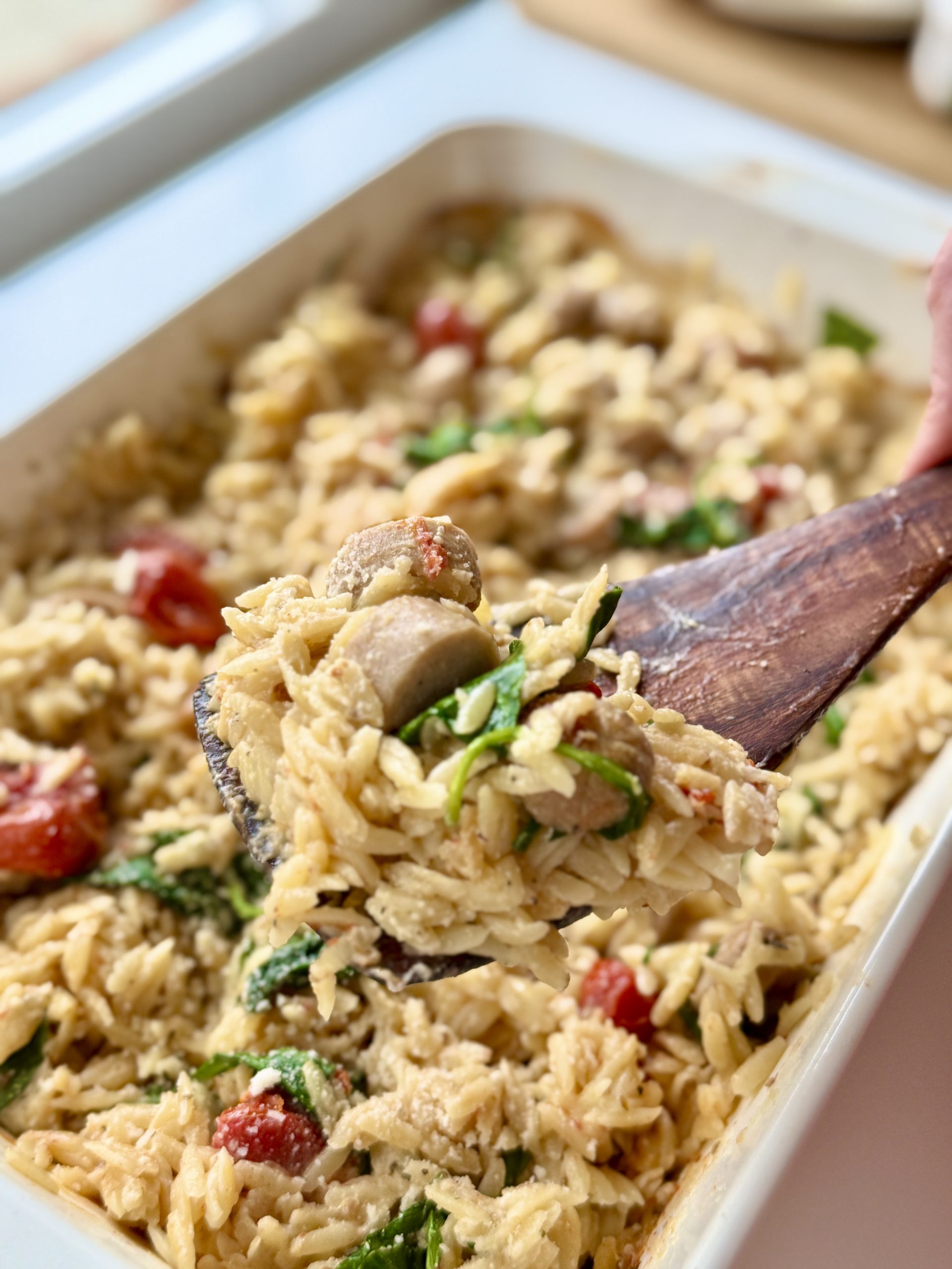 A close-up of a wooden spoon scooping creamy baked orzo pasta with mushrooms, spinach, and cherry tomatoes from a casserole dish.