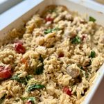 A baking dish filled with baked orzo pasta mixed with cherry tomatoes, spinach, crumbled cheese, and herbs, sitting on a kitchen counter near a window.