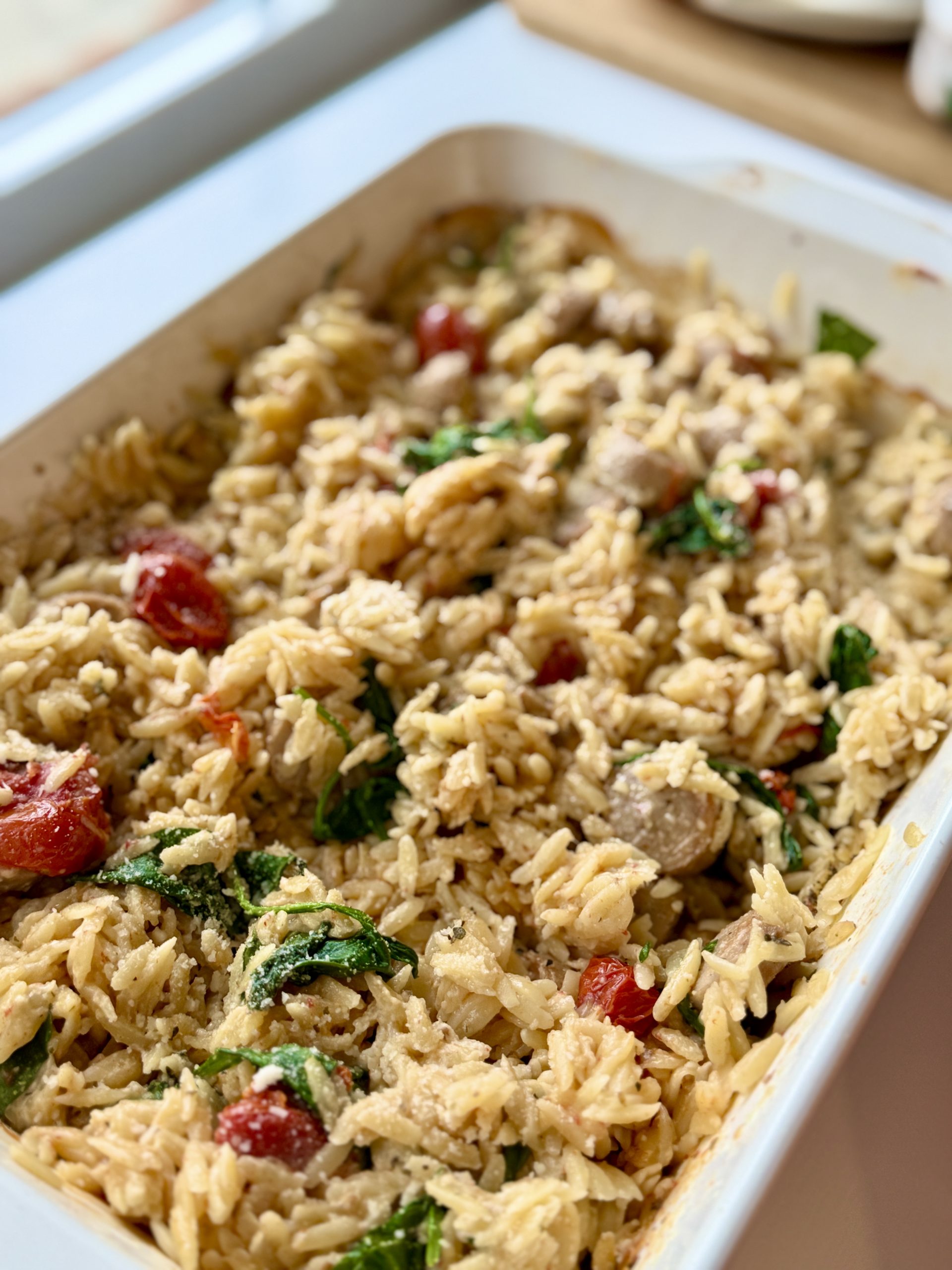 A baking dish filled with baked orzo pasta mixed with cherry tomatoes, spinach, crumbled cheese, and herbs, sitting on a kitchen counter near a window.