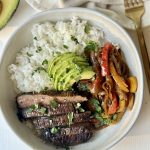 A white bowl filled with white rice, sliced grilled steak, sautéed bell peppers and onions, and sliced avocado, garnished with chopped cilantro. A gold fork and avocado half are nearby on a white surface.