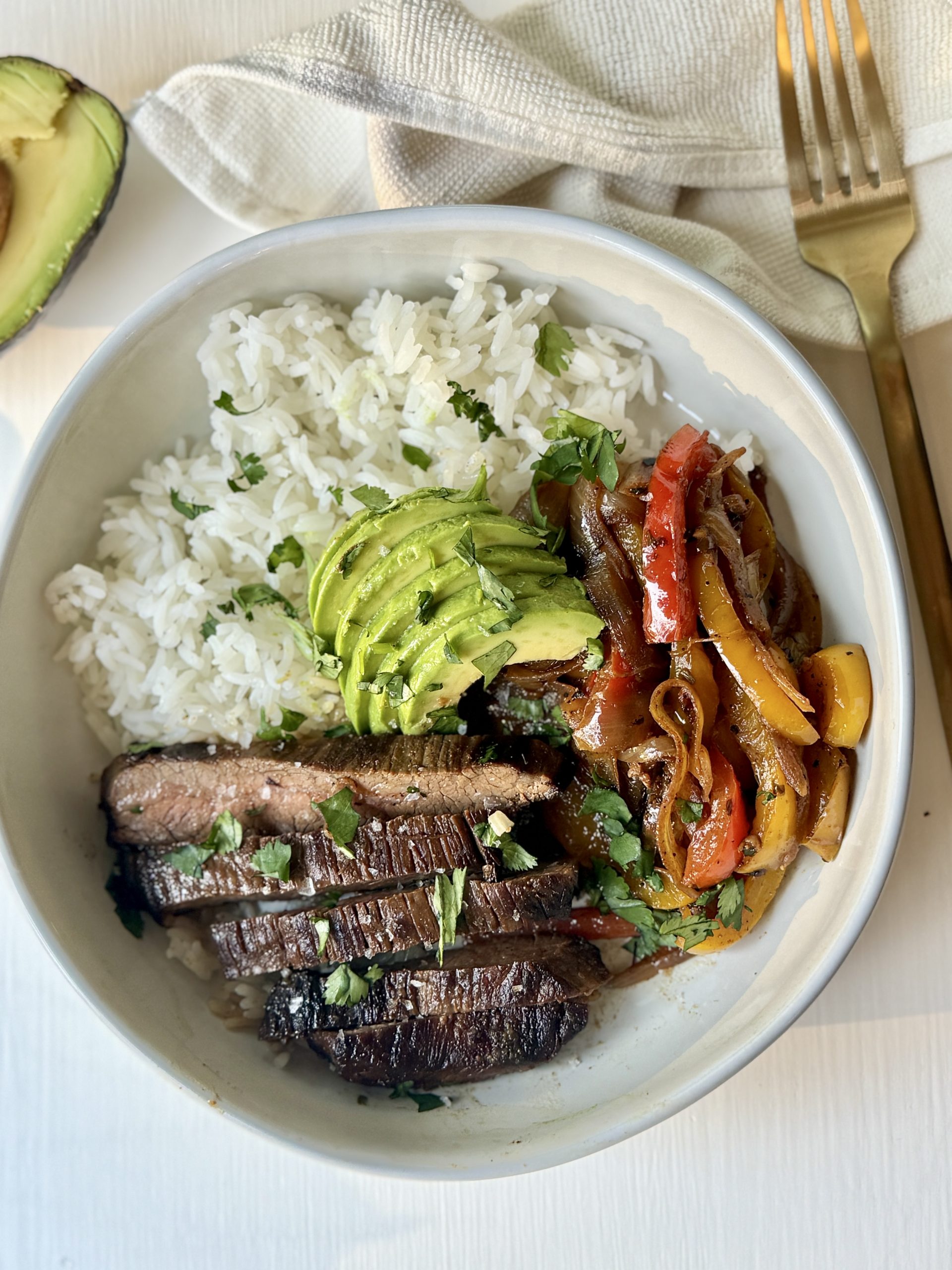 A white bowl filled with white rice, sliced grilled steak, sautéed bell peppers and onions, and sliced avocado, garnished with chopped cilantro. A gold fork and avocado half are nearby on a white surface.